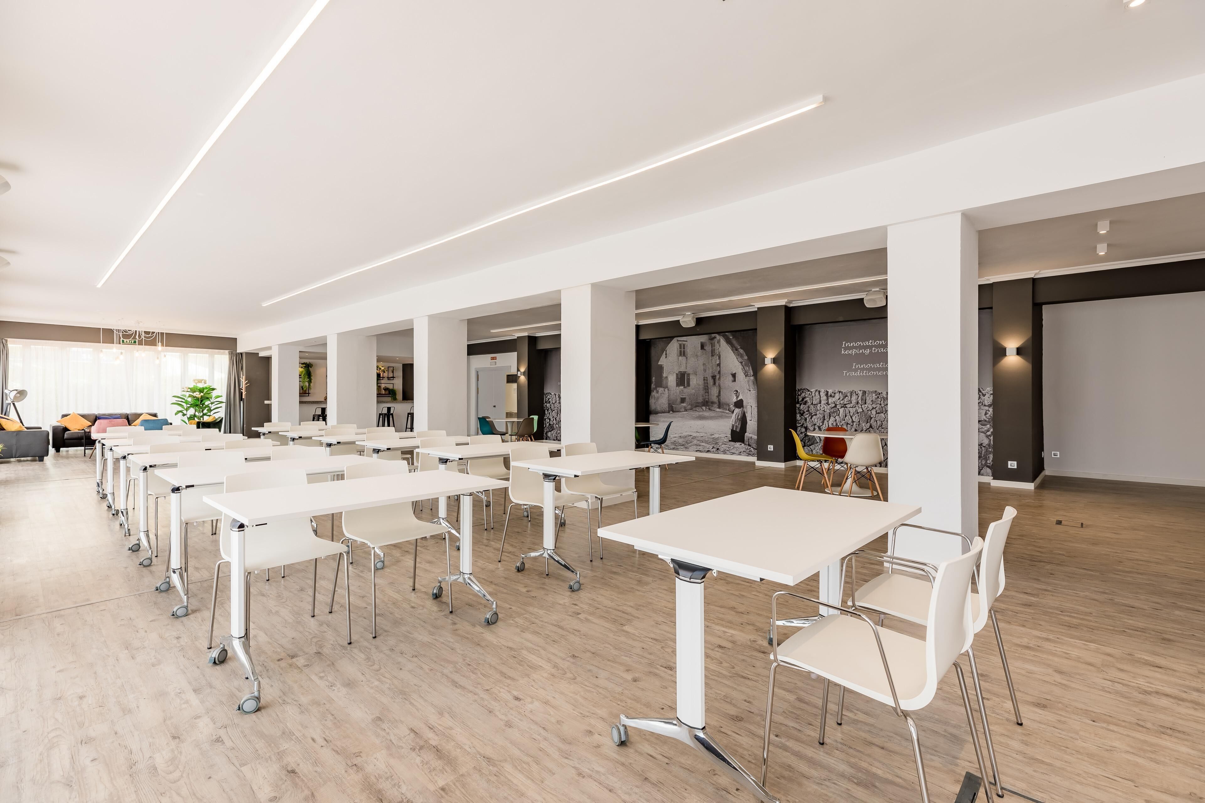 Un salón de conferencias moderno con mesas y sillas blancas, piso de madera y un mural grande en la pared.