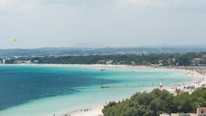 Escena de playa costera con aguas turquesas cristalinas, un muelle y vista a la ciudad en la distancia.