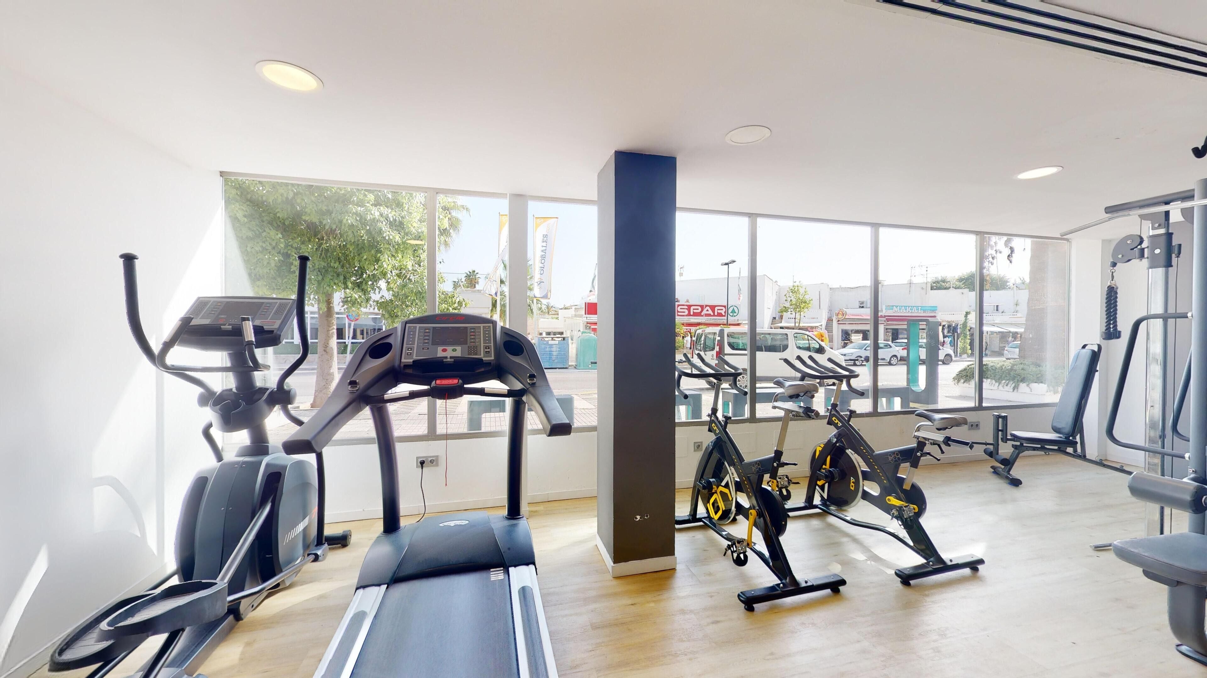 Un gimnasio con máquinas de cardio, equipo de entrenamiento con pesas y amplios ventanales que ofrecen vistas al exterior.