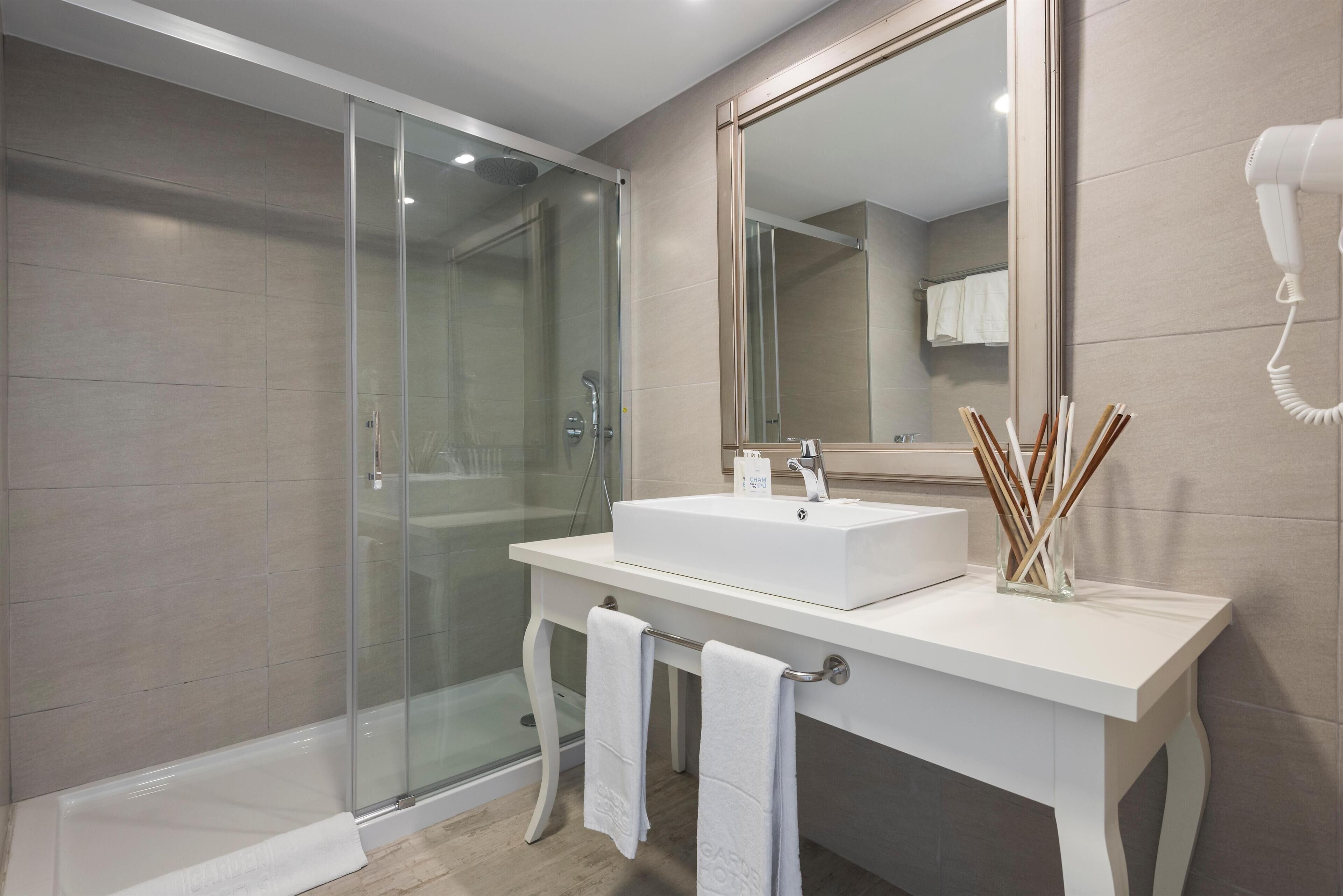 Un baño moderno con ducha acristalada, tocador blanco con un espejo grande y un secador de pelo montado en la pared.