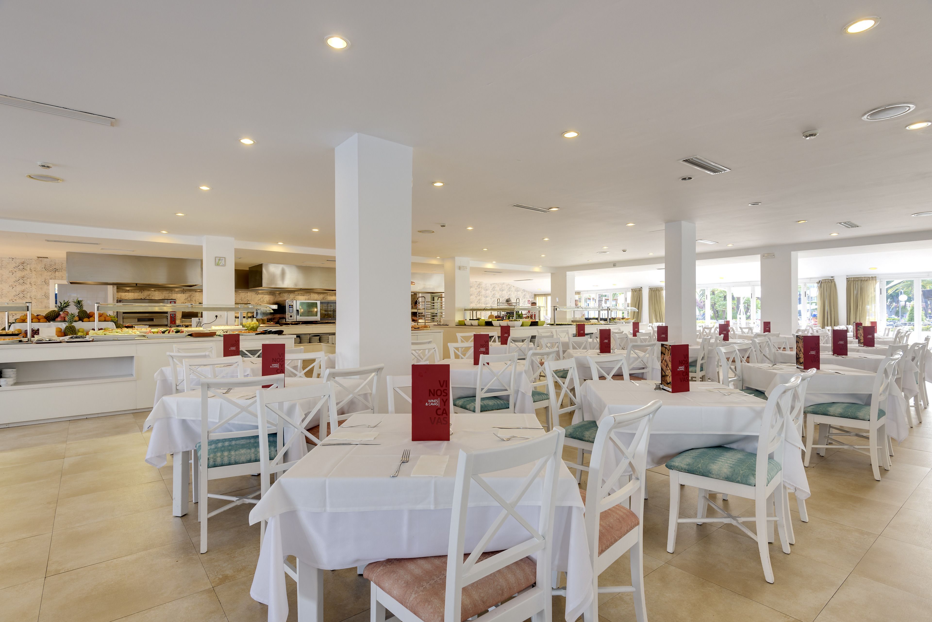 Uma sala de jantar espaçosa com mesas e cadeiras brancas, um ponto de serviço central e vista para a cozinha ao fundo.