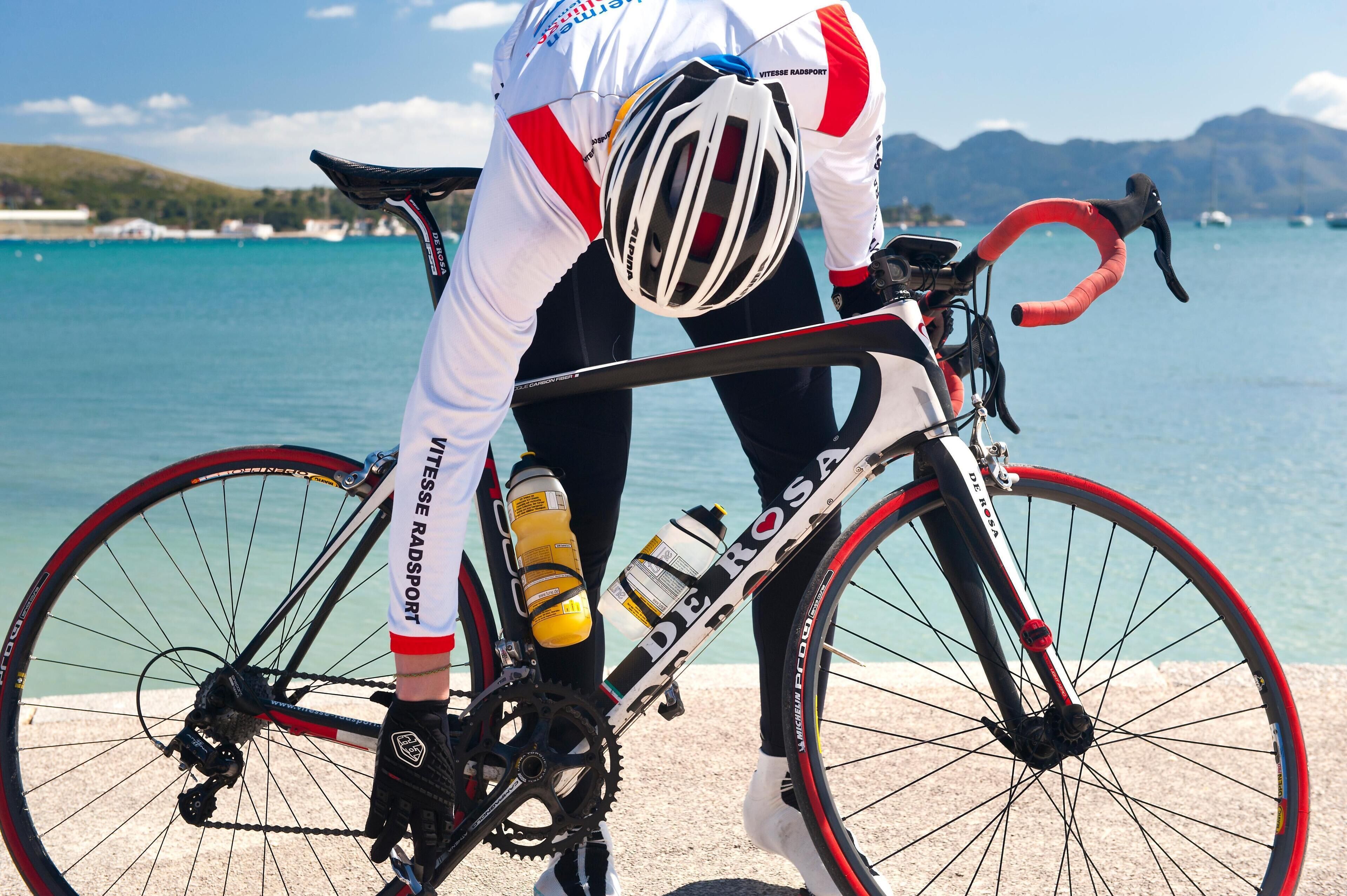 Ein Fahrrad mit einer Wasserflasche und einem Helm, platziert in der Nähe von Gewässer mit Booten und einer hügeligen Landschaft im Hintergrund.