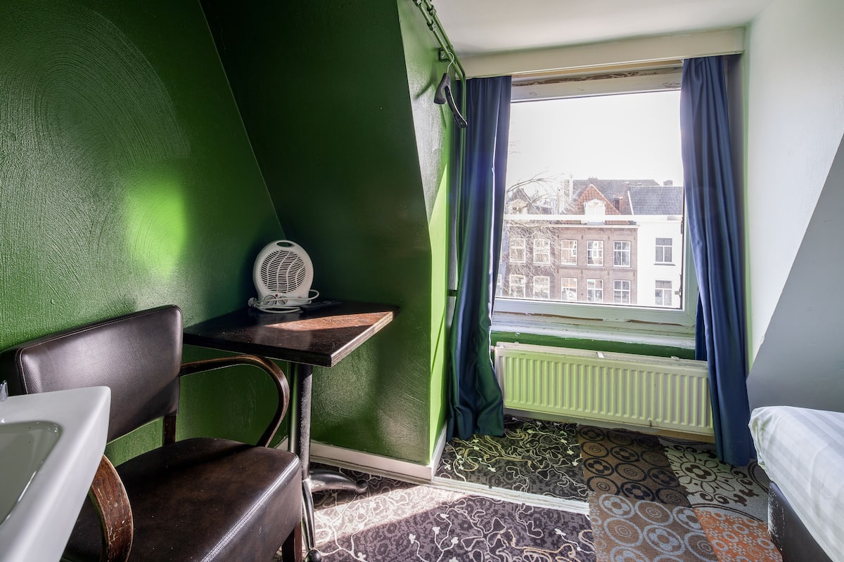 Une pièce avec un mur vert, une fenêtre avec des rideaux bleus, un radiateur, une moquette à motifs, un lit, une chaise et une petite table avec un ventilateur.