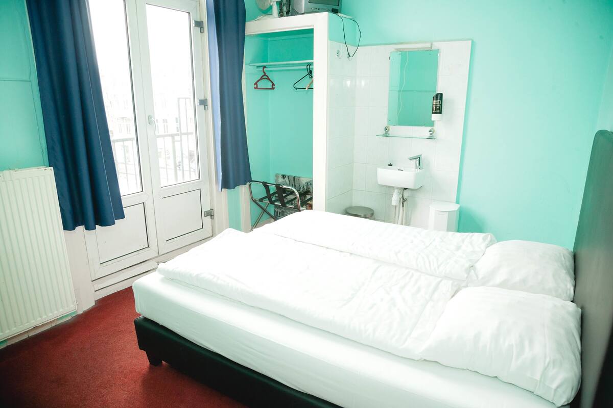 Une chambre à coucher avec un lit, une fenêtre avec des rideaux, une salle de bain avec un lavabo et un miroir, et un radiateur.