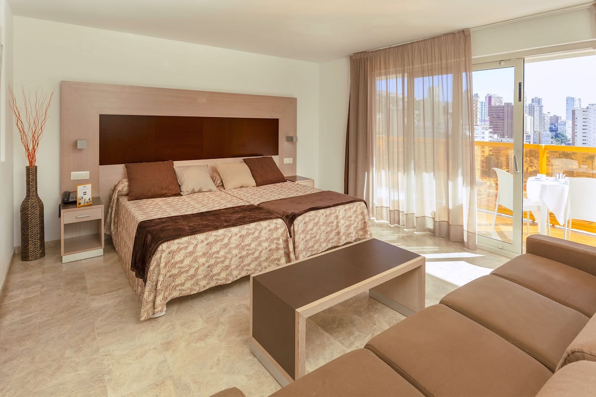 A modern hotel room with a large bed, a small bedside table, a nightstand, a sofa, and a balcony with a city view.
