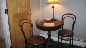 Une pièce avec une porte en bois, une table ronde surmontée d’une lampe, deux chaises et une petite table avec un bol et un livre.