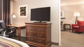 A hotel room with a bed, a wooden dresser with drawers, a desk, a television, a red chair, a lamp, and artwork on the walls.