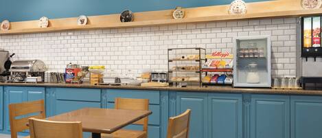 A kitchen with blue cabinets, a wooden table, and chairs. There are various kitchen appliances and a refrigerator with a beverage dispenser.