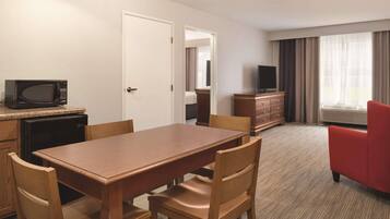 Habitación de hotel con una mesa de comedor de madera, sillas, microondas, televisor, cómoda y un sillón rojo.