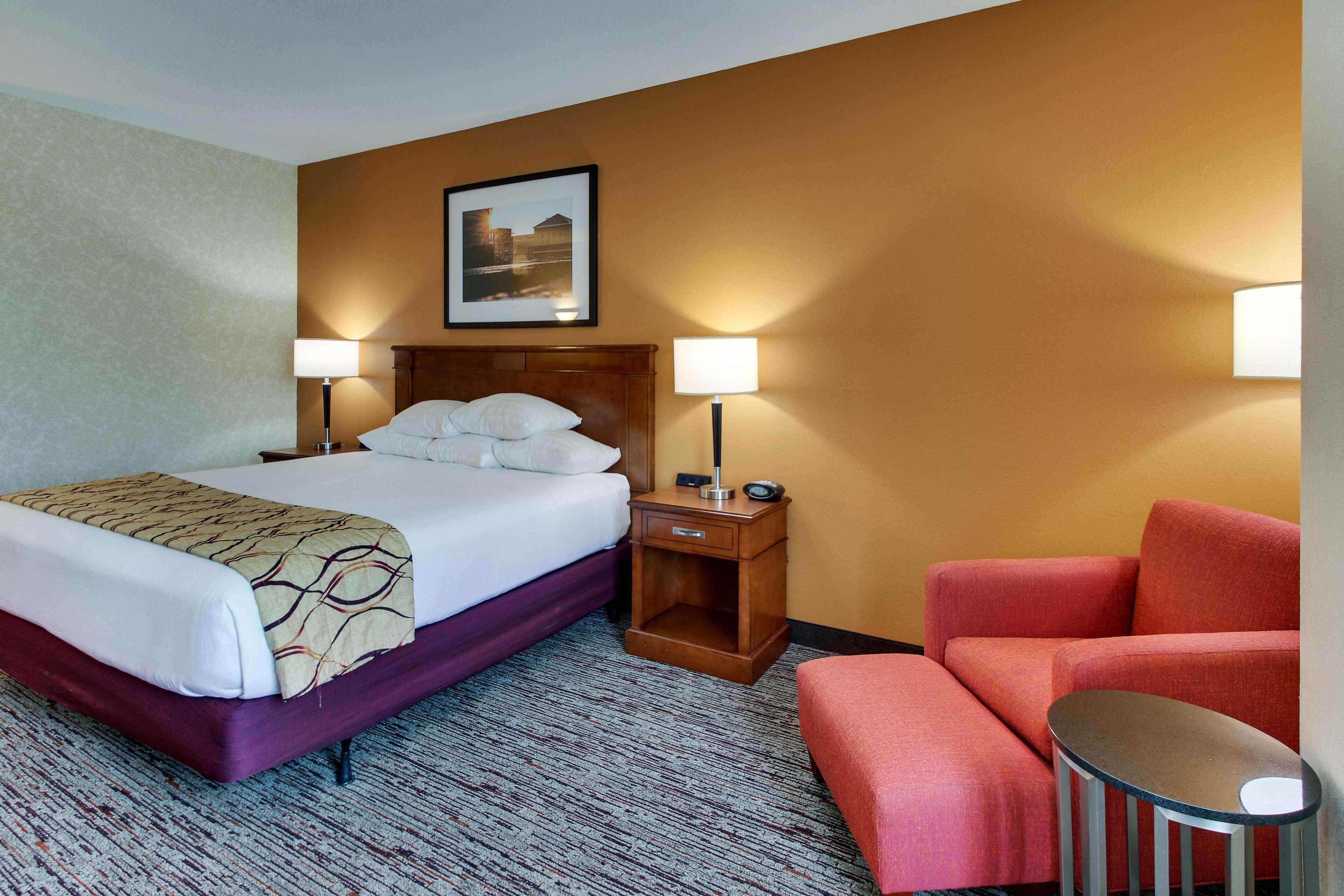 A hotel room with a bed, bedside lamps, a chair, a small table, and a framed picture on the wall.