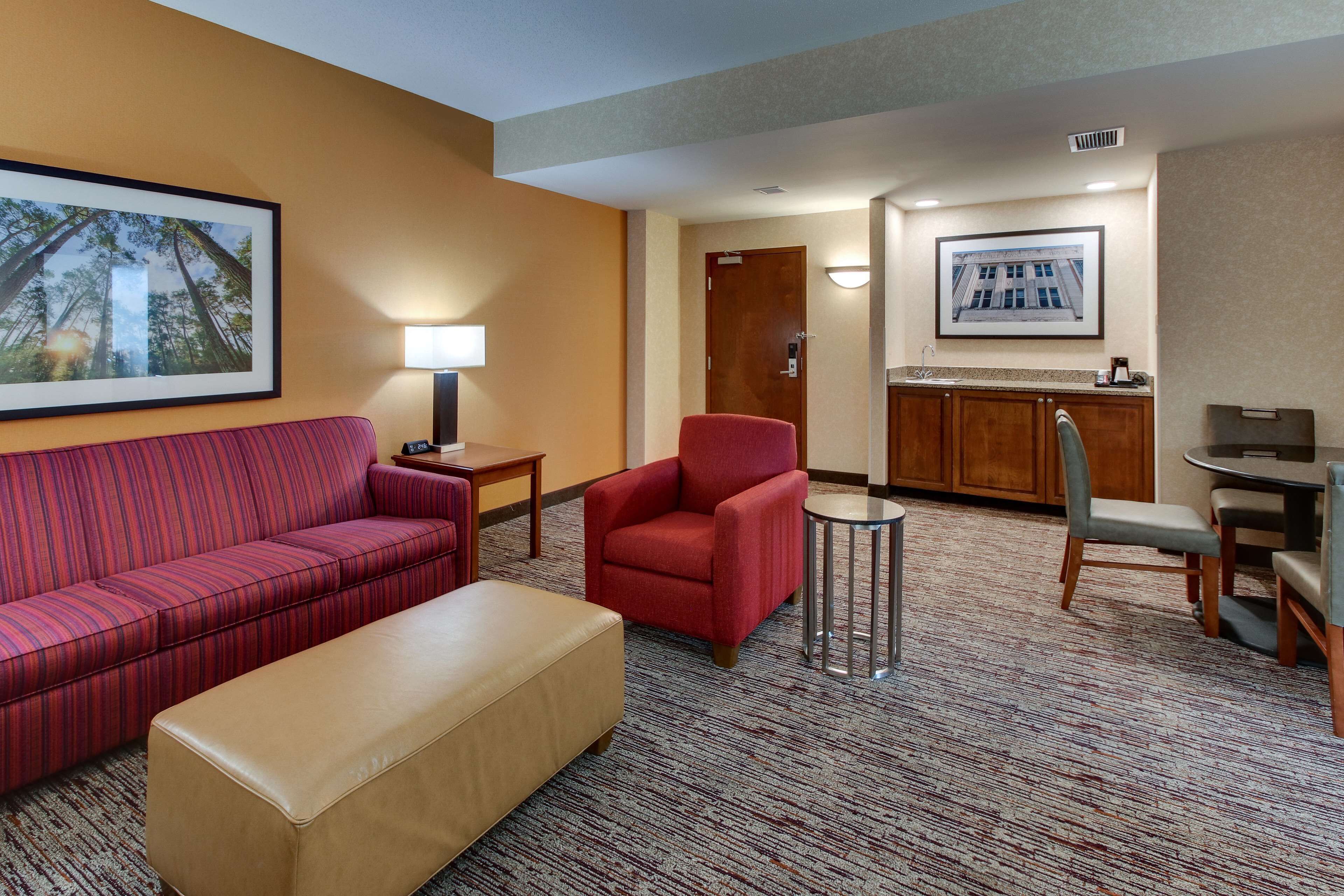 Habitación de hotel con sofá rojo, un reposapiés color beige, un sillón rojo, una mesita redonda y una zona de kitchenette con pileta y encimera.