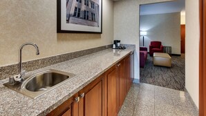 A hotel kitchen with a stainless steel sink, granite countertop, and wooden cabinets. There is a framed picture on the wall and a red chair in the background. - Drury Inn & Suites Meridian (Meridian)