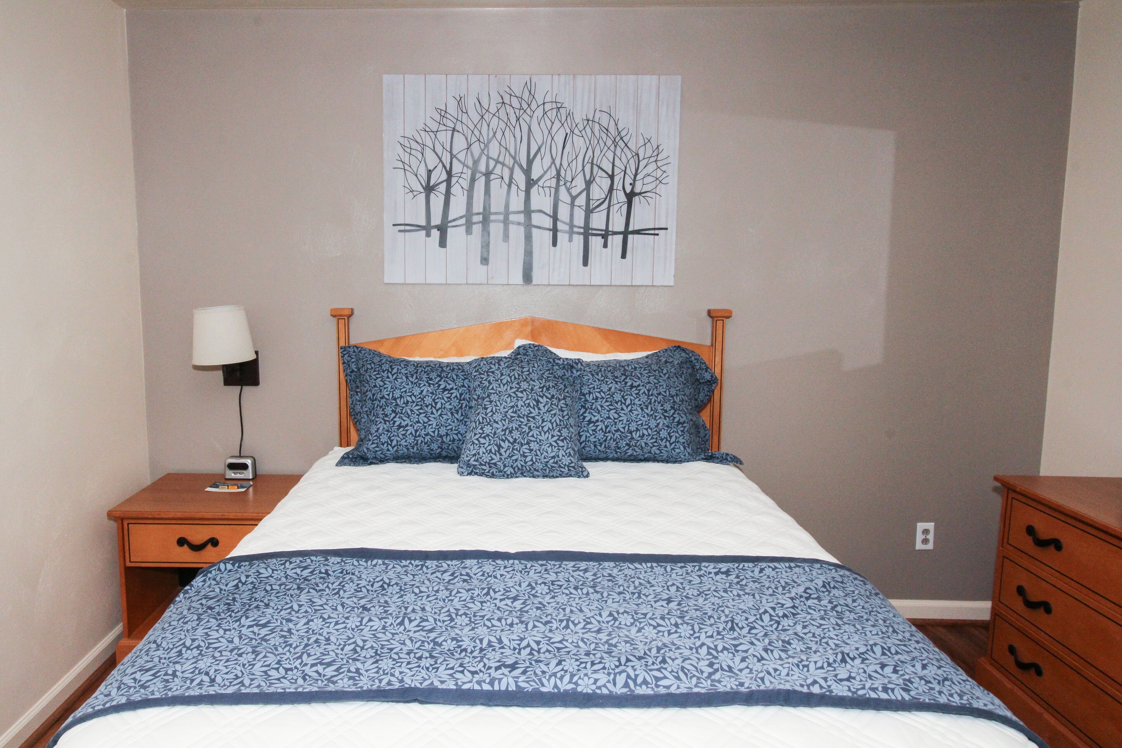 A neatly made bed with a blue and white patterned bedspread, a wooden headboard, and a wall-mounted painting of trees.