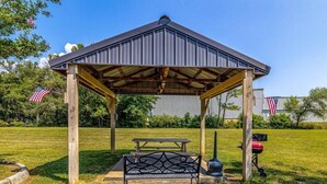 Un kiosque doté d’un toit en métal et de poutres en bois, une table de pique-nique et un barbecue, le tout installé dans un espace verdoyant orné de drapeaux américains.