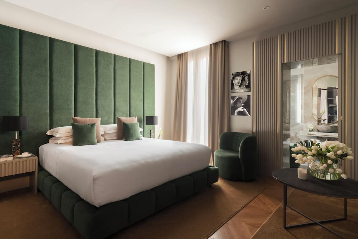 A modern hotel room with a large bed, a green upholstered headboard, a small round table with a vase of flowers, and a seating area with a green armchair.