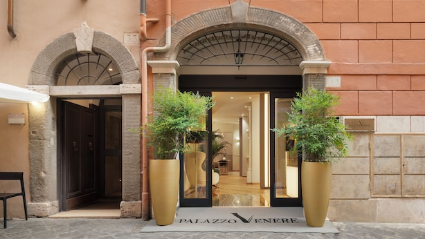 Entrada de un hotel con fachada de piedra, dos plantas grandes en macetas y un letrero que dice "Palazzo Veneri".