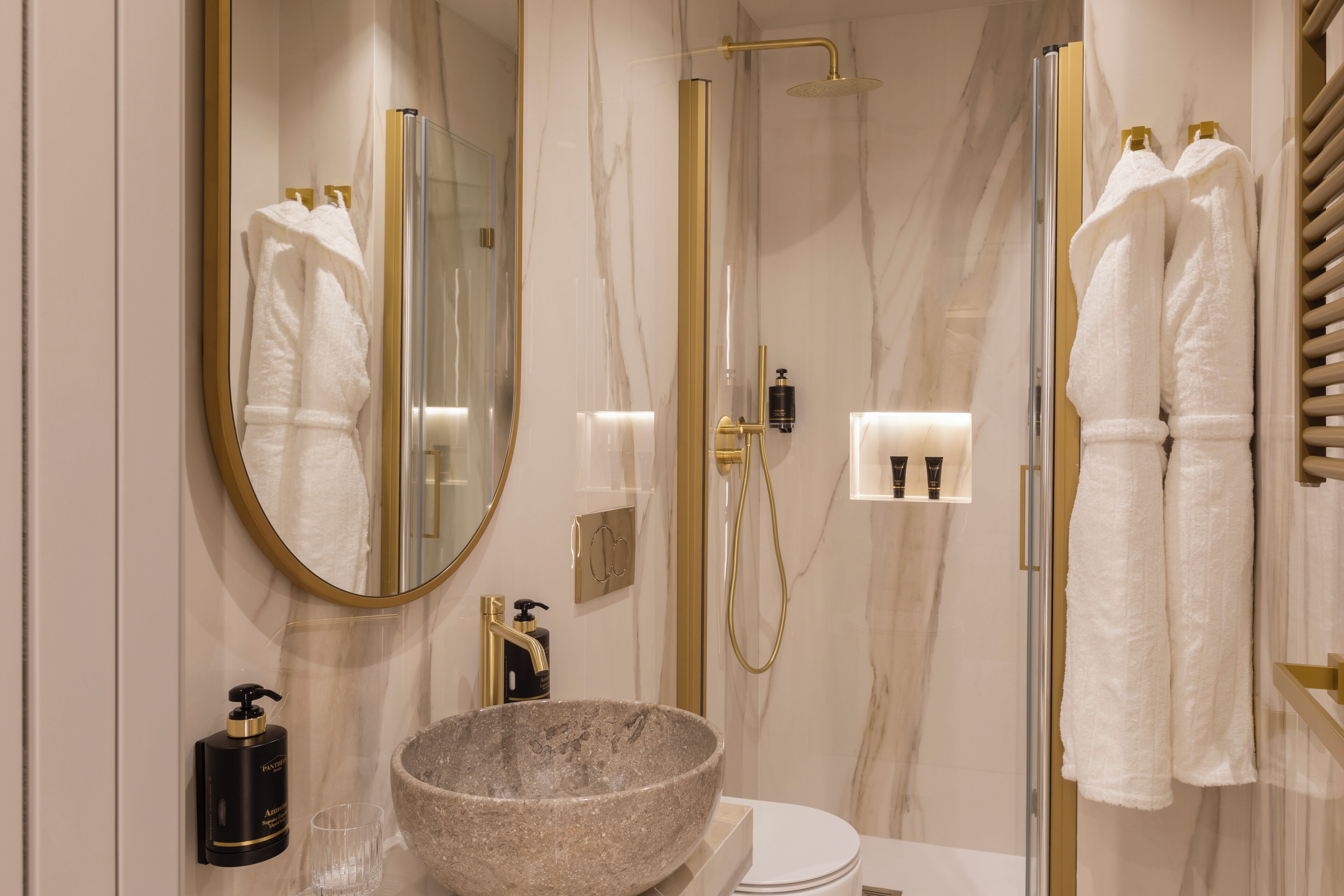 A modern bathroom with a marble wall, a large oval mirror, a stone sink, a gold showerhead, and white towels.