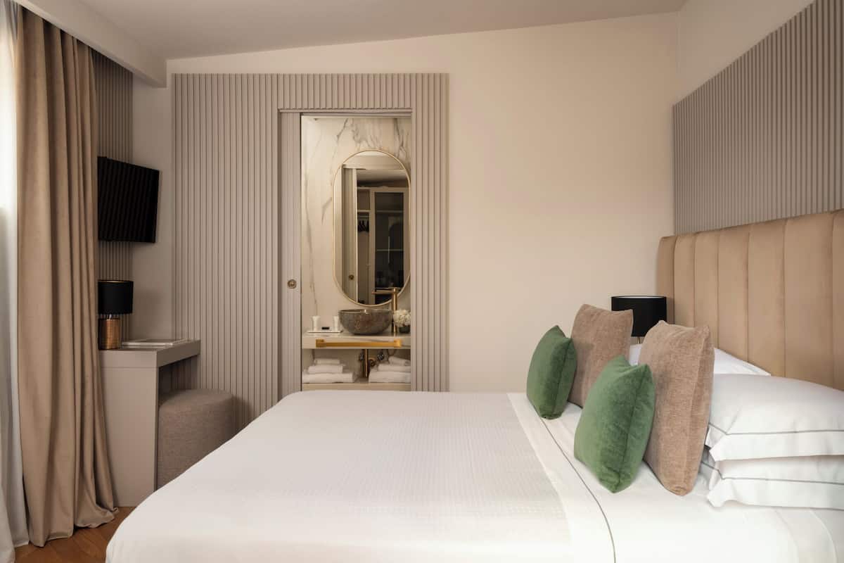 A hotel room with a bed, a bedside table, a TV, a mirror, and a bathroom visible through an open door.