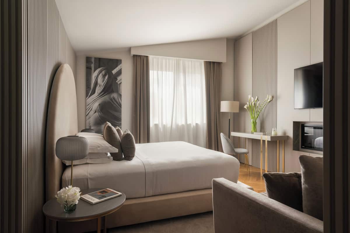 A modern hotel room with a large bed, a desk, a chair, a TV, and a window with curtains.