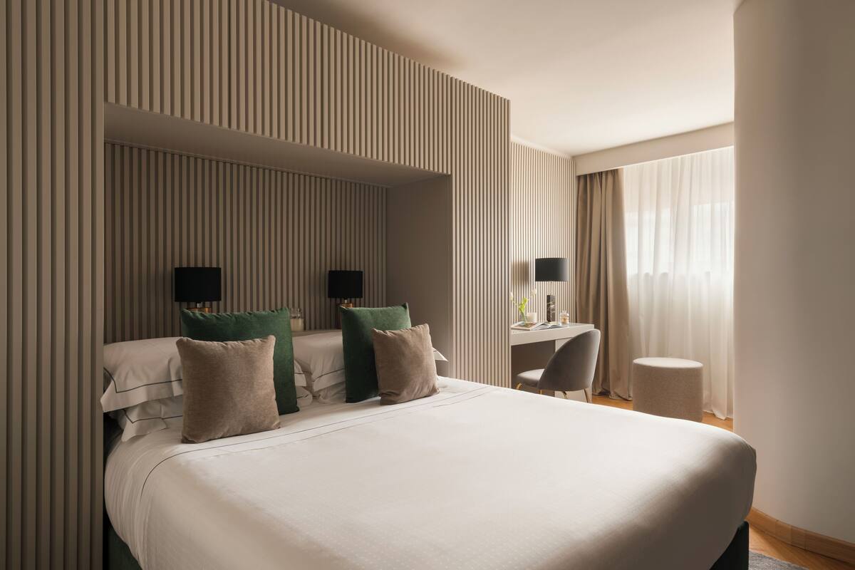 A modern hotel room with a large bed, two bedside tables with lamps, a desk, and a chair.