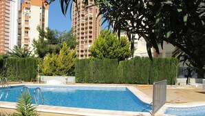 A swimming pool with a metal fence, surrounded by greenery and tall buildings in the background.