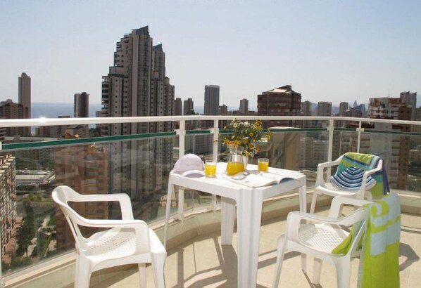 Un balcón con mesa y sillas, un florero con flores y vistas a la ciudad.
