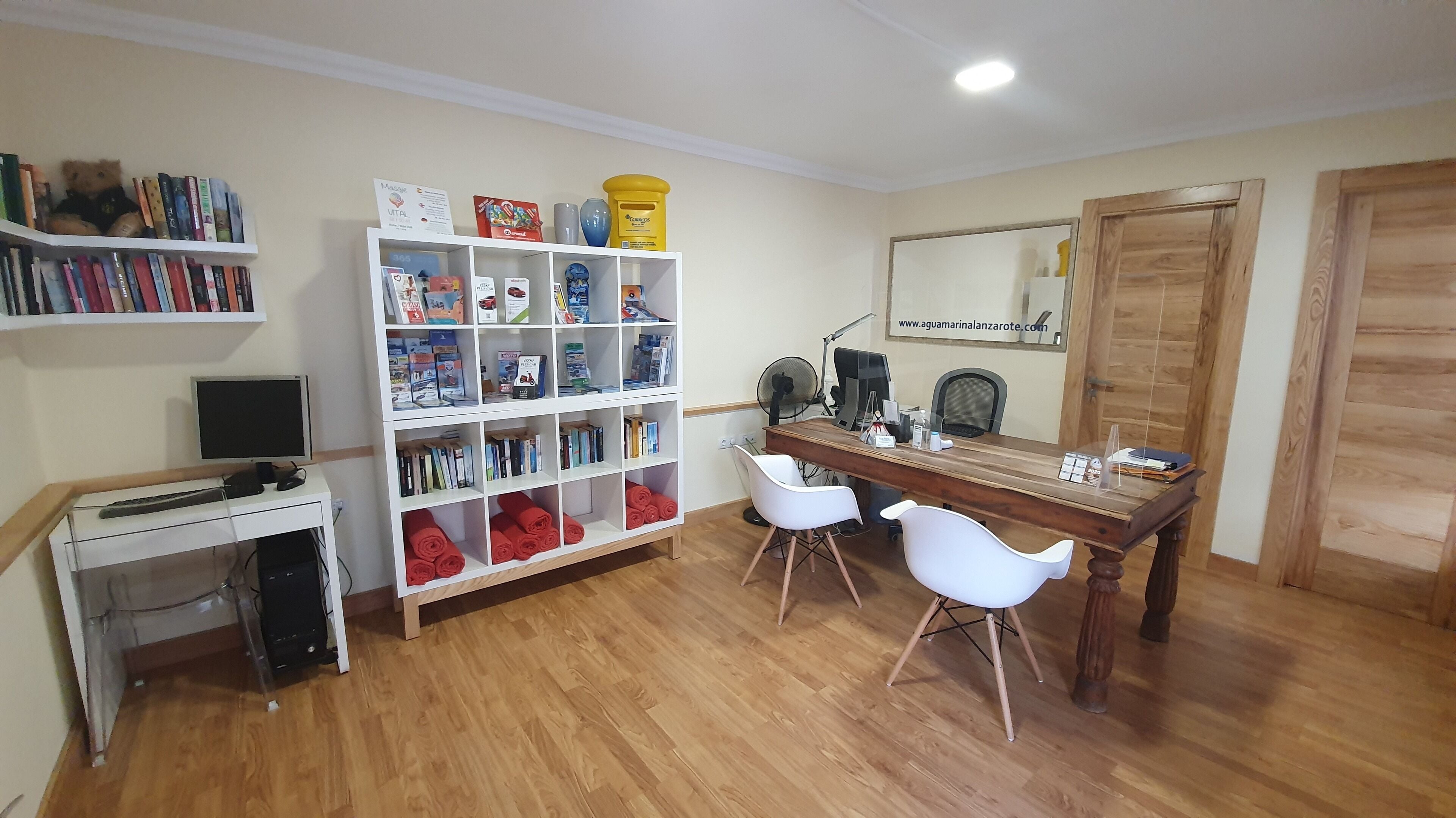 A room with a wooden desk, two white chairs, a bookshelf filled with books and magazines, a computer monitor, and a mirror on the wall.