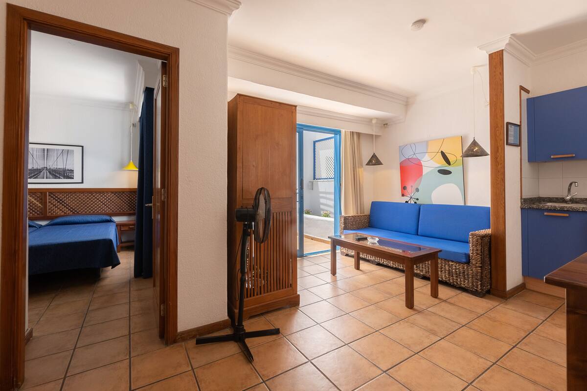 Habitación de hotel con cama, sofá azul, una mesita, ventilador y zona de cocina con armarios azules.