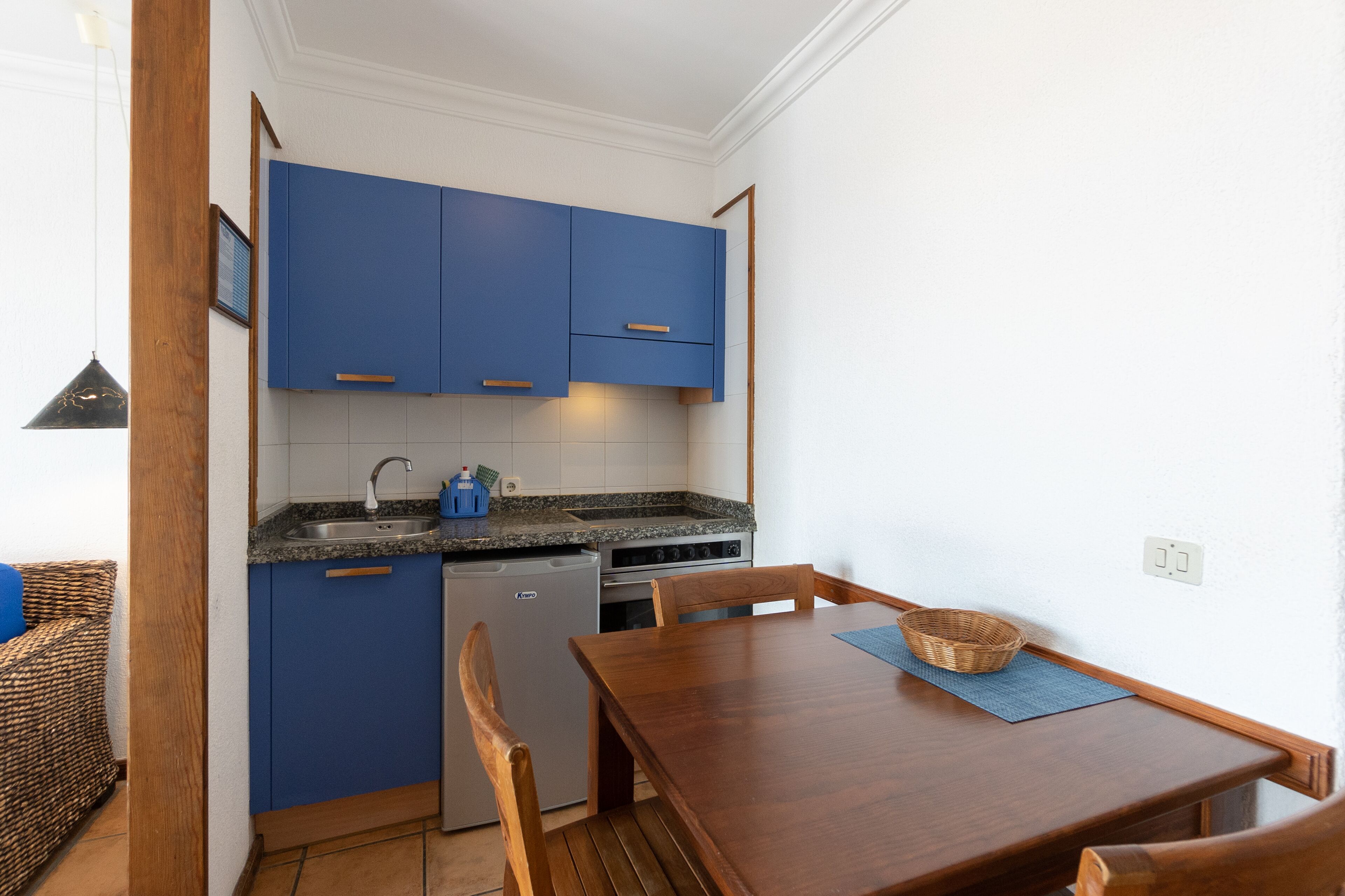 A compact kitchen with blue cabinets, a built-in dishwasher, and a dining area with a wooden table and chairs.