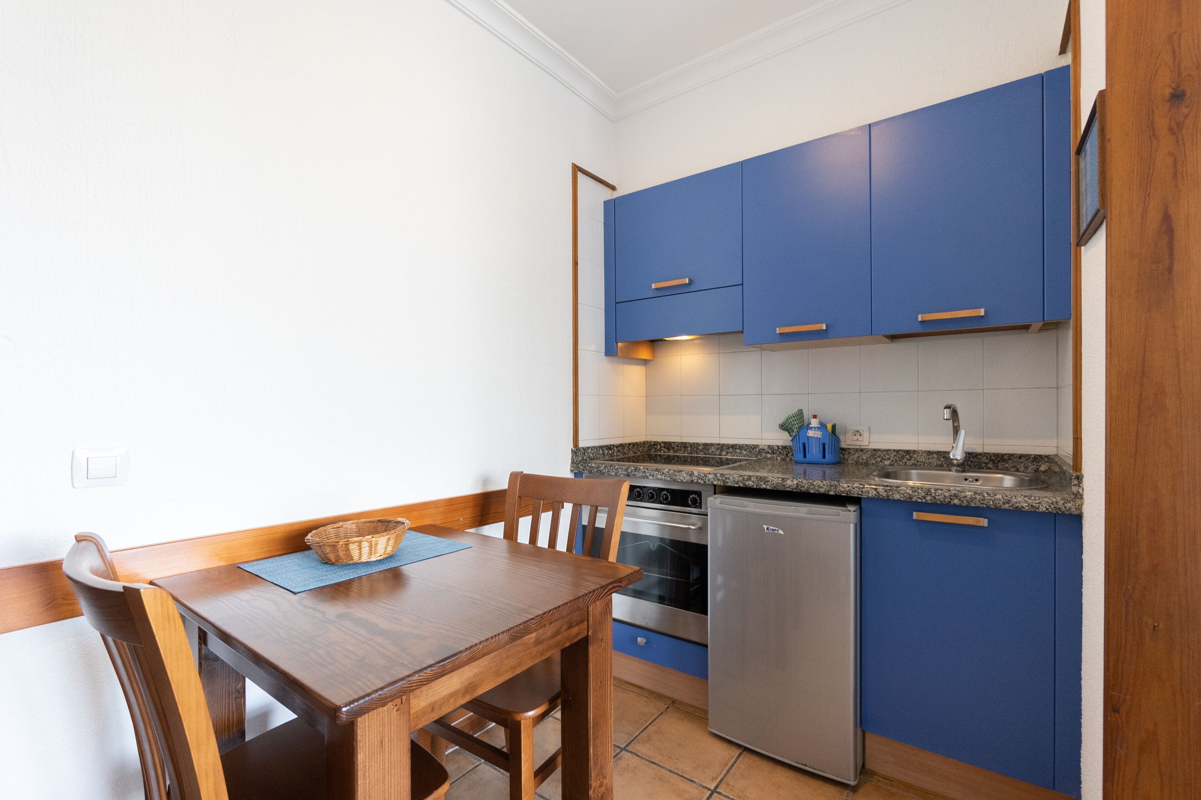 A compact kitchen with blue cabinets, a stainless steel refrigerator, a built-in oven, and a granite countertop.