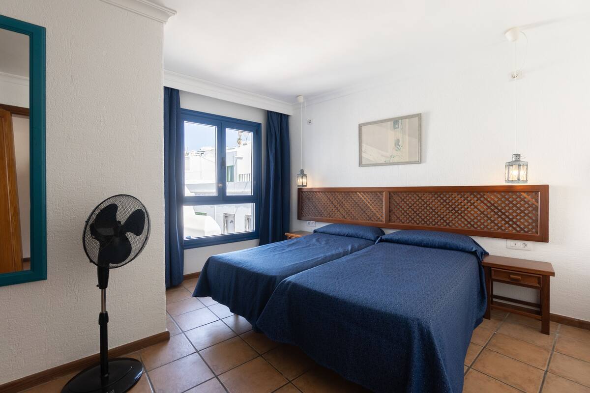 Una habitación de hotel con dos camas, un ventilador de pie, una ventana con cortinas, una mesita de noche y una lámpara fijada a la pared.