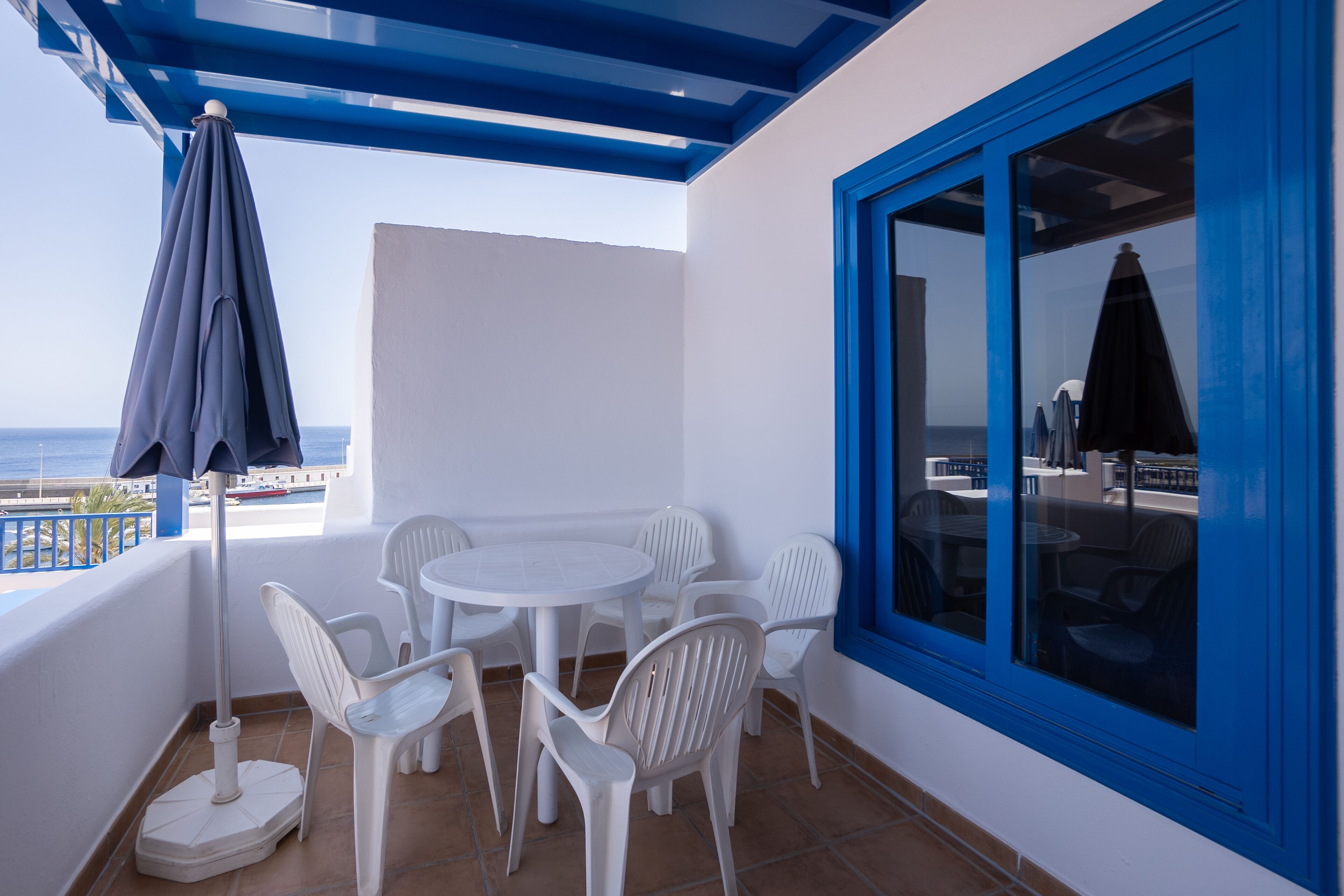 Una terraza con sillas de plástico blanco, una mesa, un paraguas azul y vistas al mar.