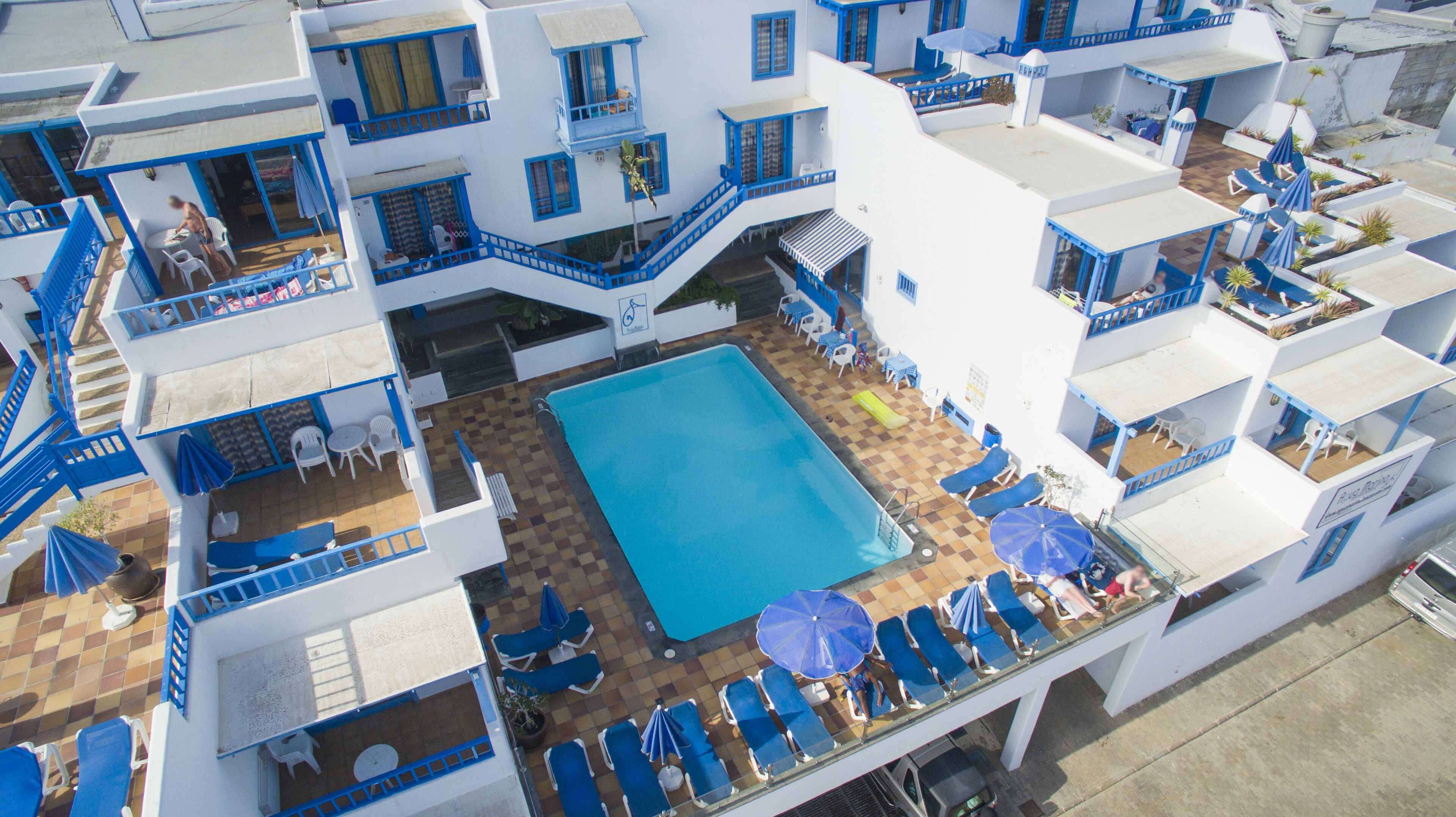 Un hotel con piscina central rodeada de sillones azules y sombrillas, paredes blancas y detalles en azul.