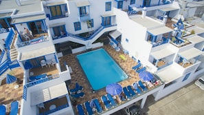 A hotel with a central swimming pool surrounded by blue lounge chairs and umbrellas, white walls, and blue accents.