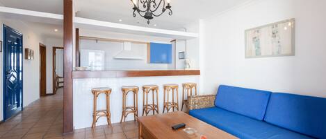 A modern living room with a wooden coffee table, a blue sofa, and a kitchen area with a bar stool.