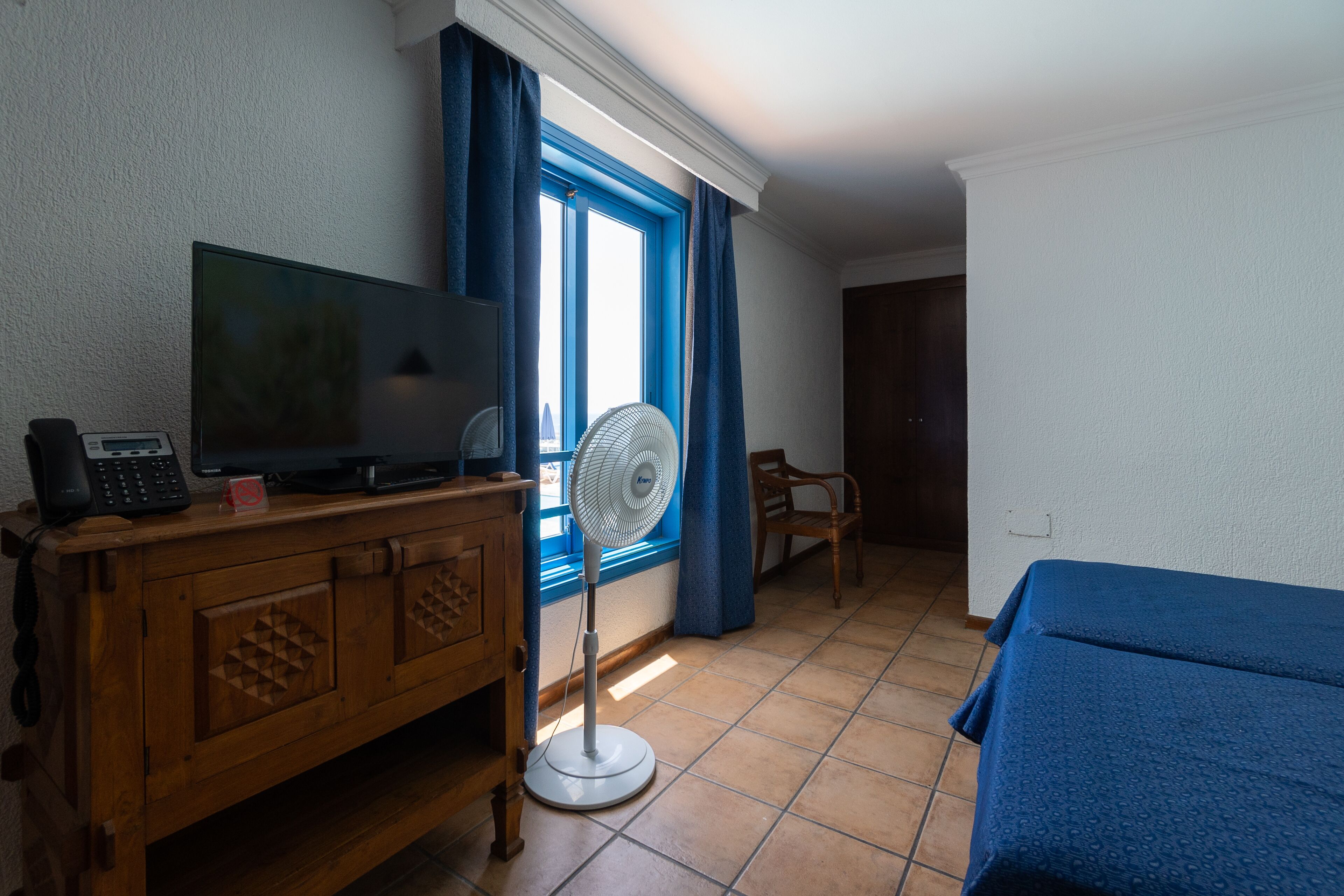 A room with a wooden cabinet, a television, a standing fan, a window with blue curtains, and a bed with a blue bedspread.