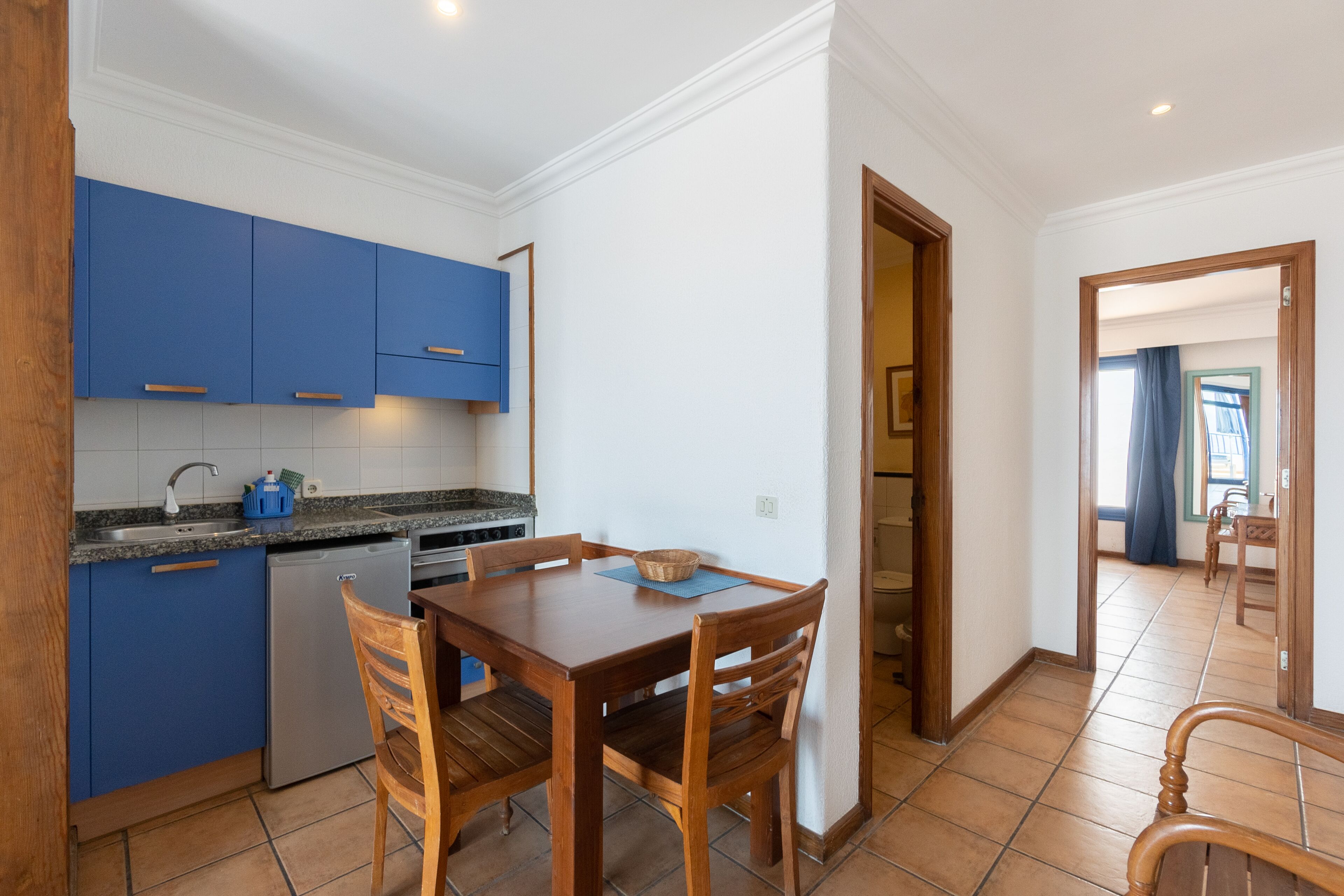 A compact kitchen with blue cabinets, a small refrigerator, and a dining table with chairs.