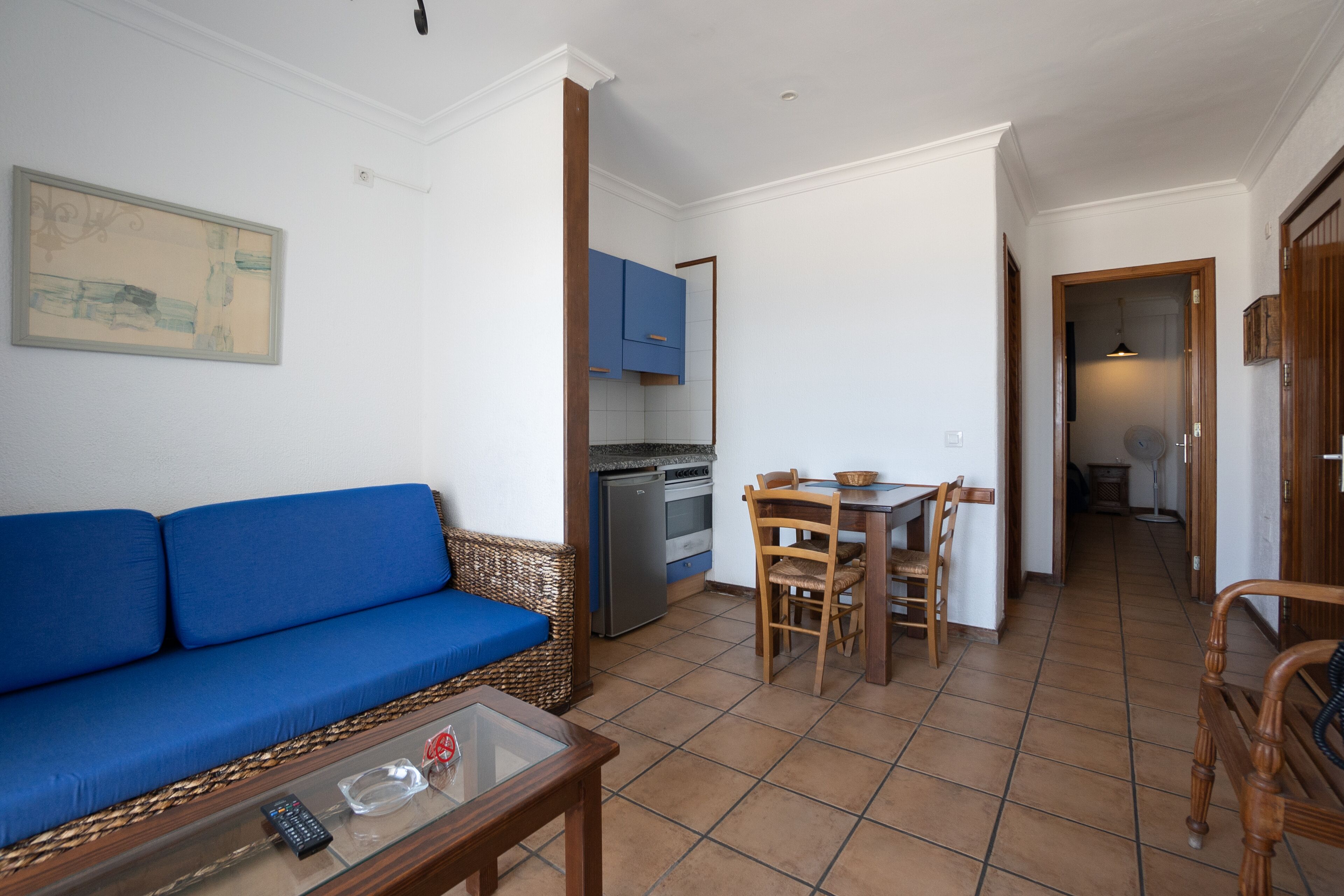 A living room with a blue sofa, a glass coffee table, and a kitchen area with cabinets and a dining table.