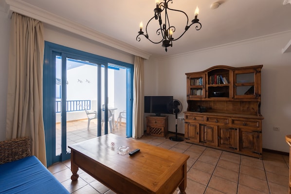A room with a wooden table, a TV, a bookshelf, and a balcony with a view.