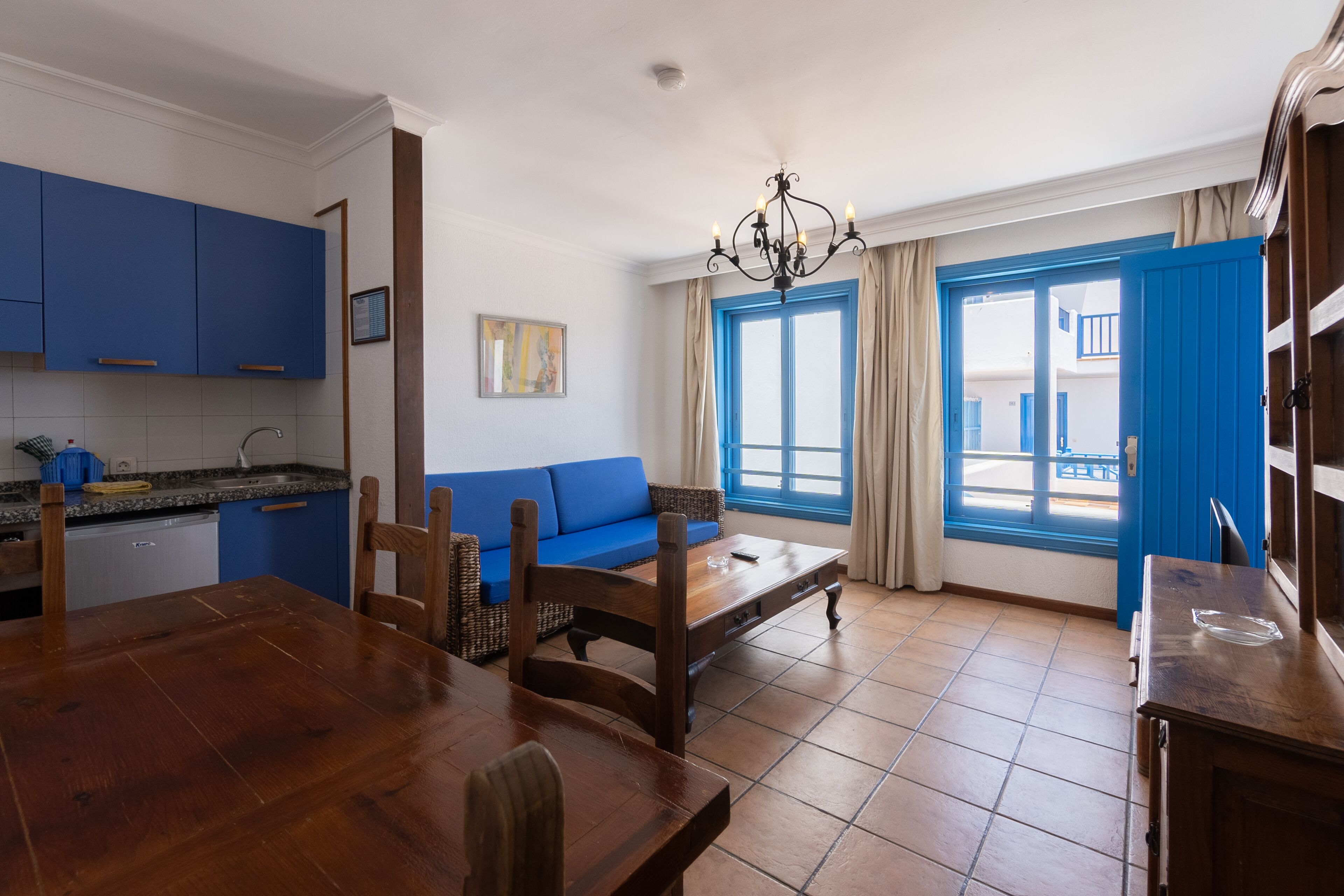 A room with a dining table, chairs, a sofa, a chandelier, and a view through a window with blue shutters.