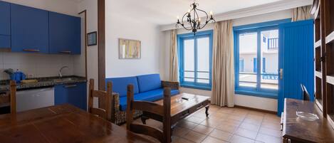 A room with a dining table, chairs, a sofa, a chandelier, and a view through a window with blue shutters.