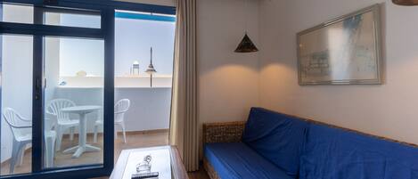 Quarto com sofá azul, mesa de centro de vidro, cadeira de vime e vista para uma varanda com cadeira branca e luminária.