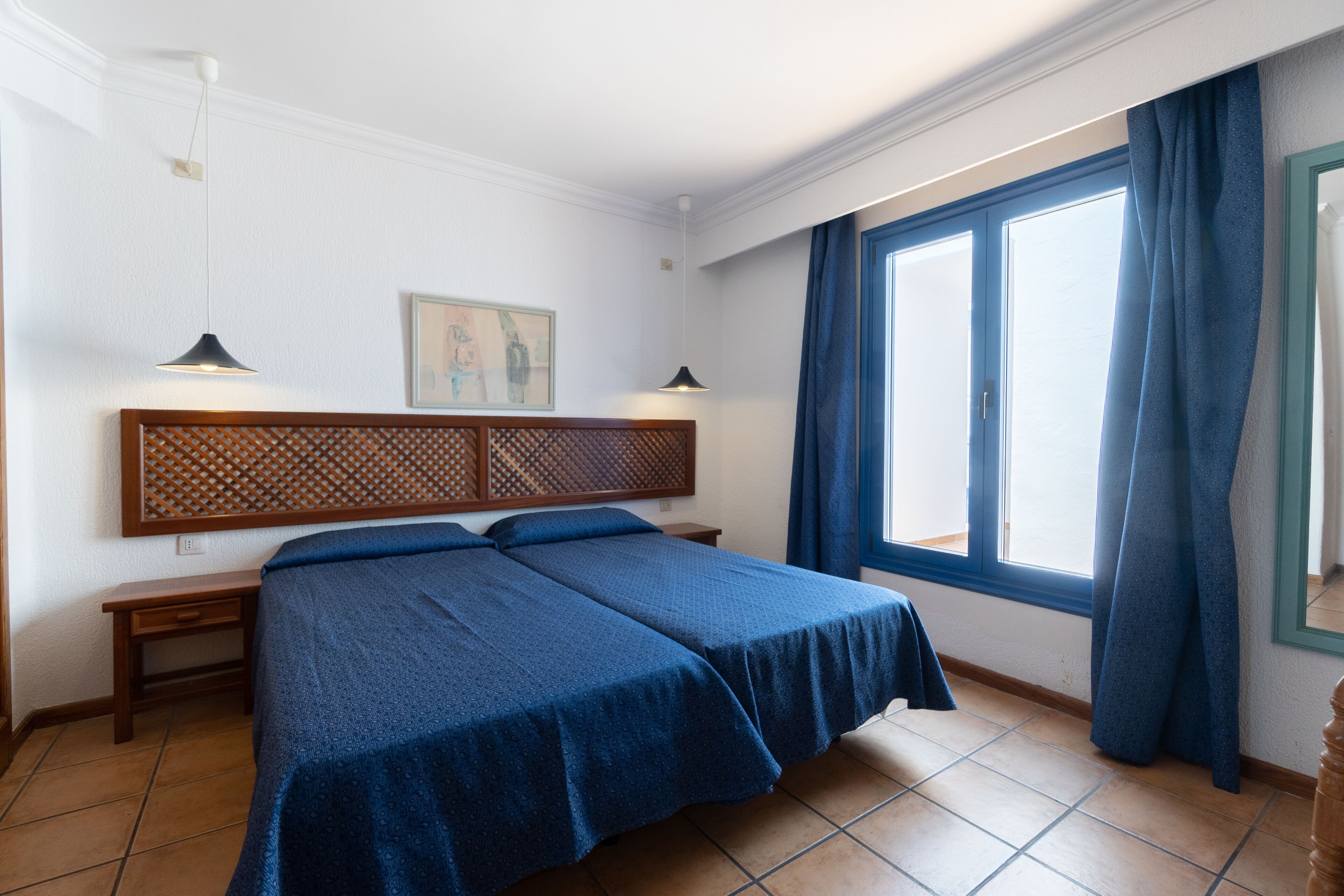 A double bed with a wooden headboard, two bedside tables, a window with blue curtains, and a wall-mounted picture.