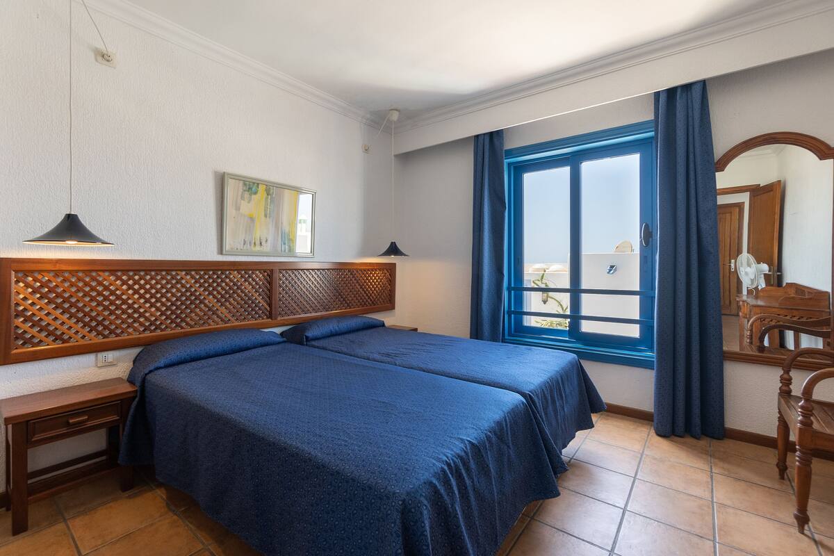 Habitación de hotel con dos camas, cabecero de madera, escritorio, silla, ventana con cortinas y un cuadro en la pared.