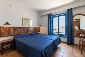 A hotel room with two beds, a wooden headboard, a desk, a chair, a window with curtains, and a painting on the wall.