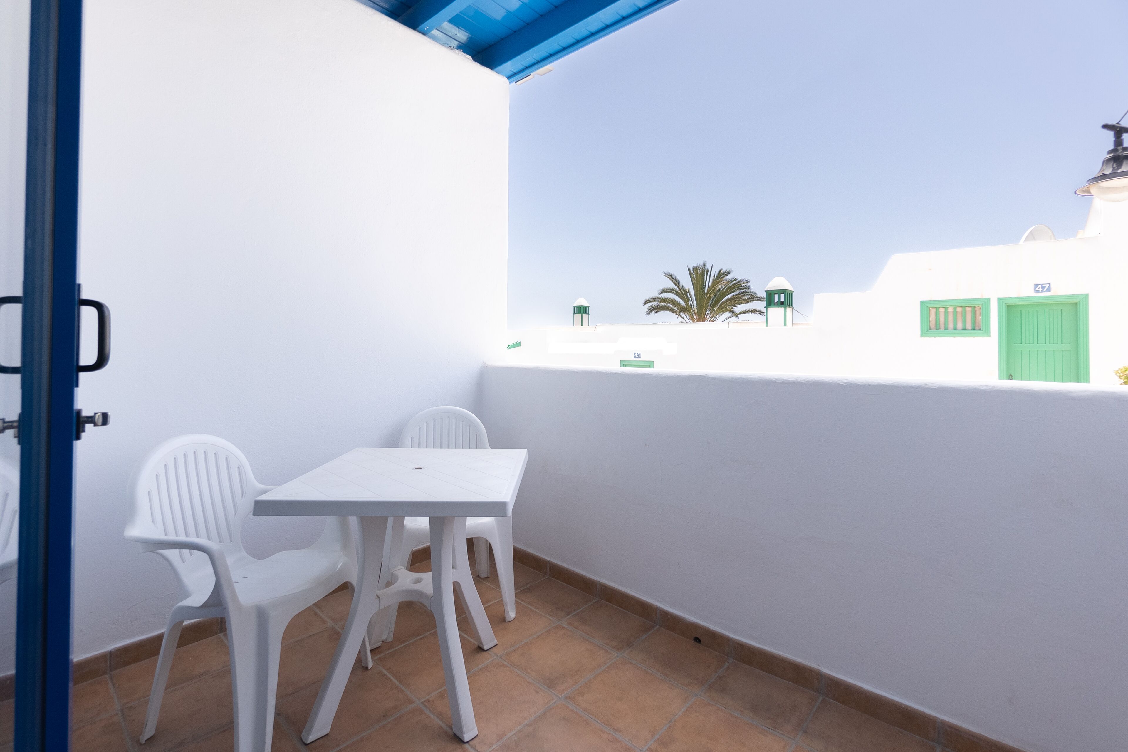 A small outdoor balcony with a white table and two white chairs, a blue door, and a view of a building with green doors and a palm tree.