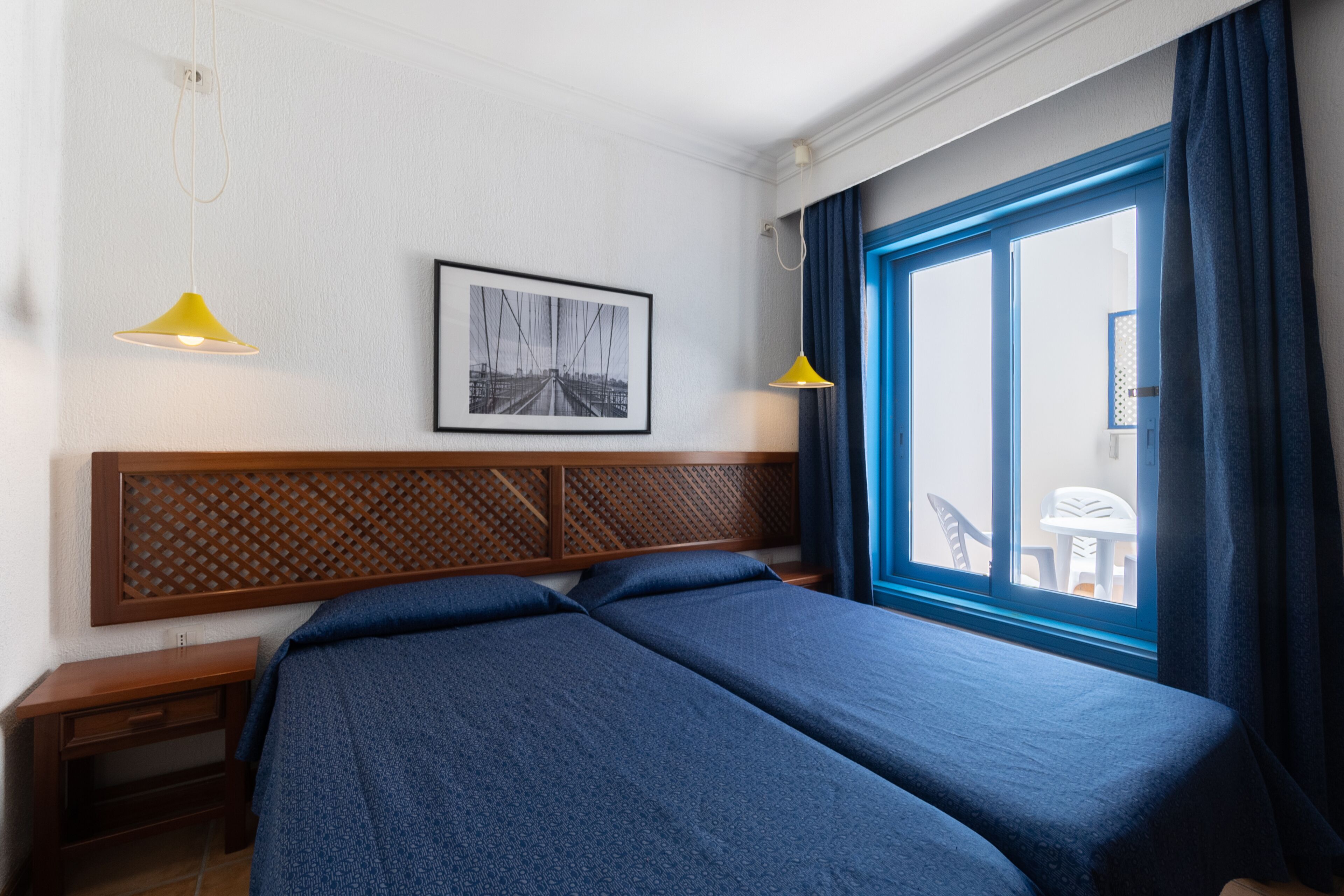 Habitación de hotel con una cama grande, cabecera con diseño de celosía, un cuadro en la pared y vistas al exterior a través de una ventana con cortinas azules.