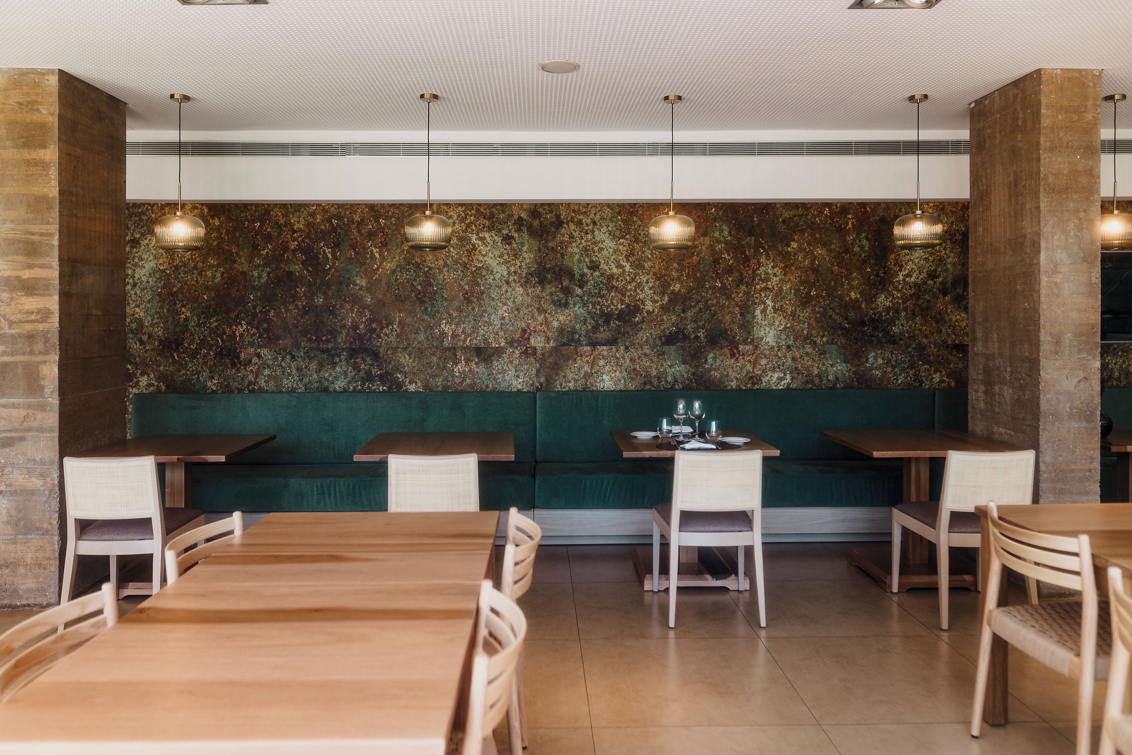 A restaurant interior with wooden tables and chairs, green cushioned seating, and pendant lighting.