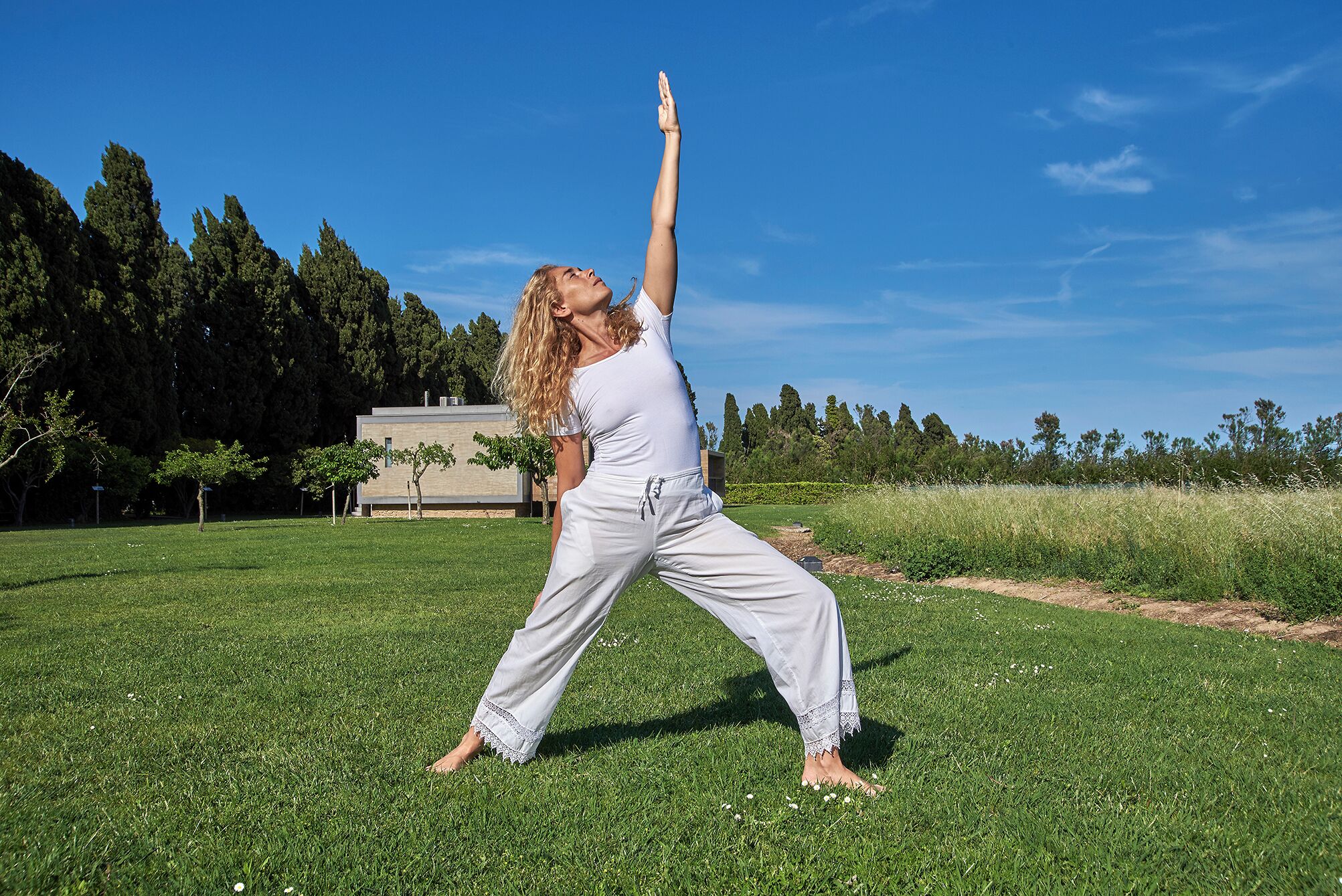Uma pessoa fazendo uma postura de yoga em área gramada ao ar livre, com árvores e um prédio ao fundo.