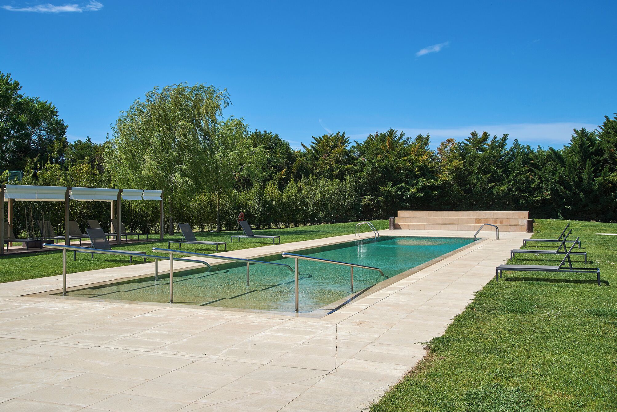 A rectangular swimming pool with a clear blue water, surrounded by a paved deck and grassy areas.